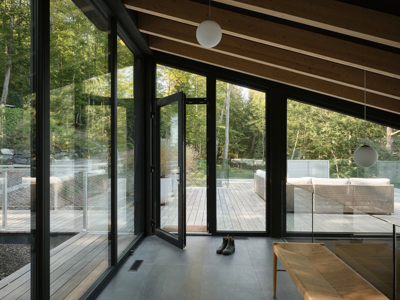 Black-framed folding glass doors opening from concrete floor to timber deck with glass railing in wooded setting