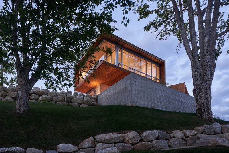 Illuminated glass pavilion cantilevering over terraced lawn with stone walls and deciduous trees at dusk