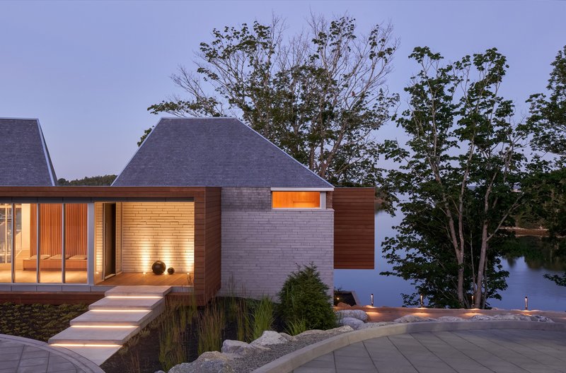 Illuminated pavilion volumes with brick and timber cladding beside a lake at twilight