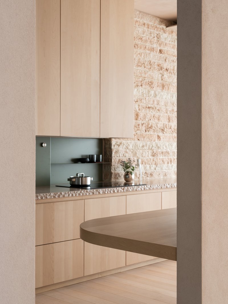 View through doorway into kitchen with pale timber cabinetry, terrazzo counter and exposed brick wall