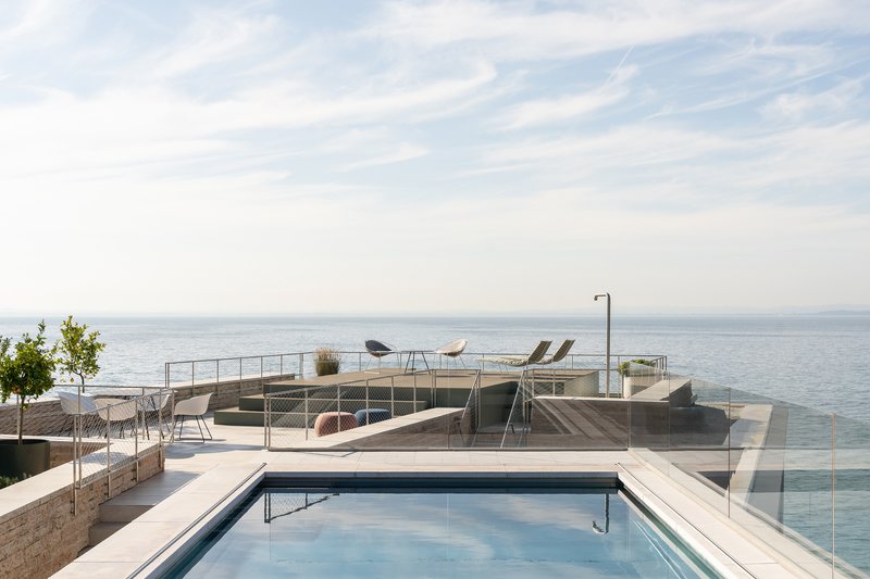 Rooftop terrace with swimming pool, glass balustrade and timber decking facing open water under soft clouds