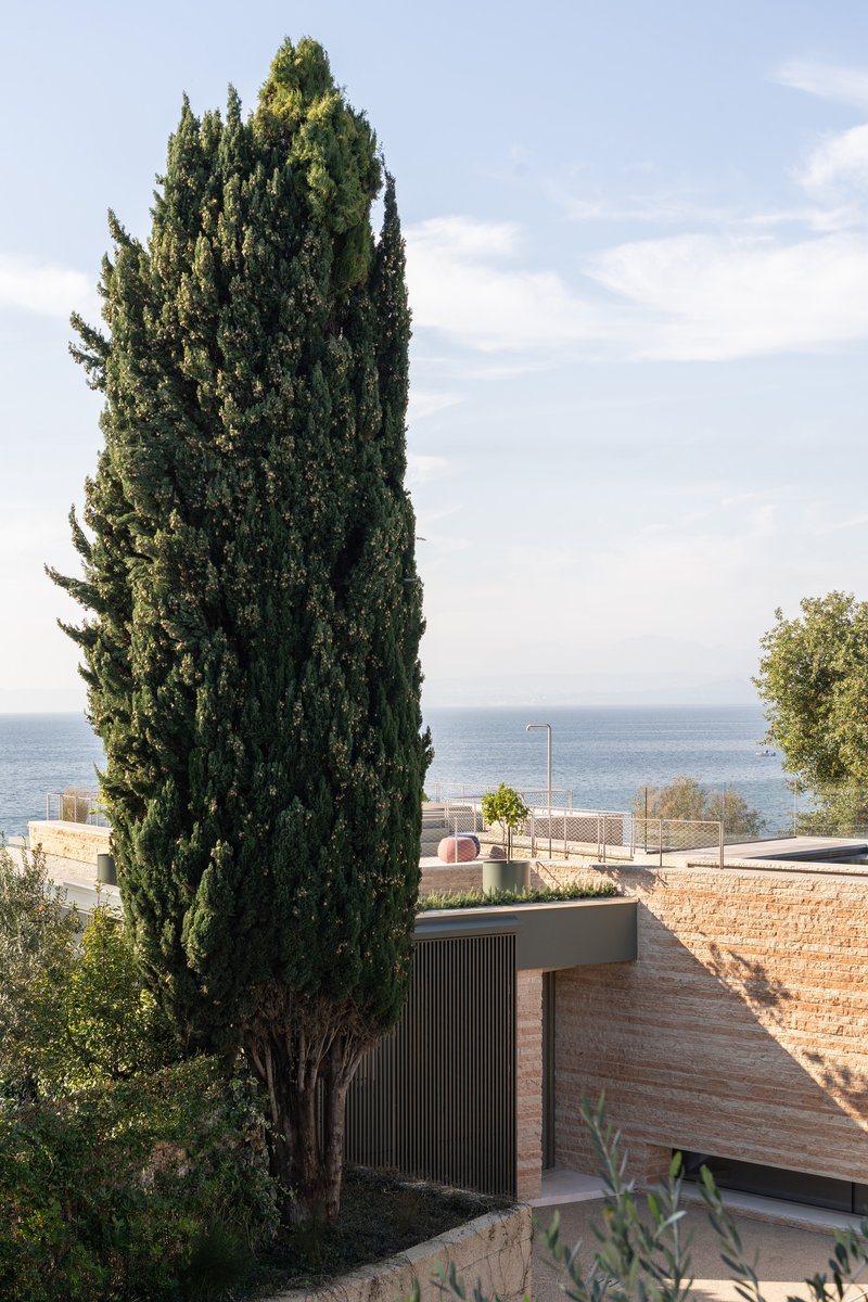 Terraced roofline with stone walls and vertical timber cladding beside a tall cypress tree overlooking the sea