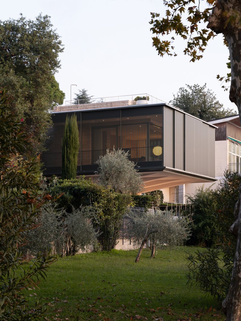 Two-storey facade with vertical screening and timber deck seen through olive trees and garden