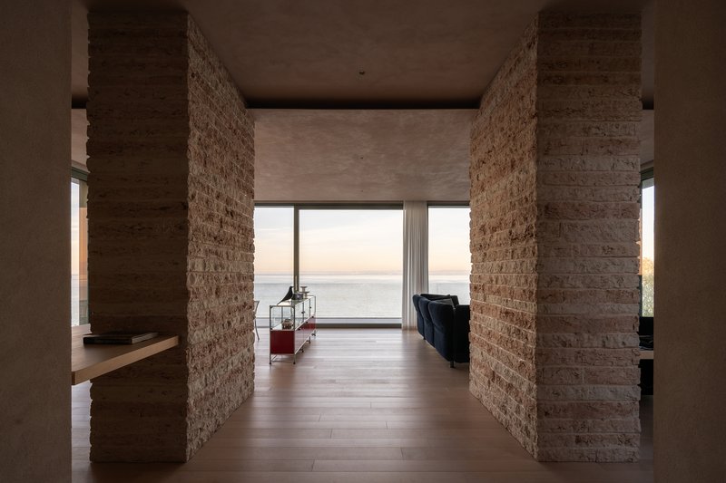 Interior hallway framed by rough stone columns leading to a glazed opening with ocean views at dusk