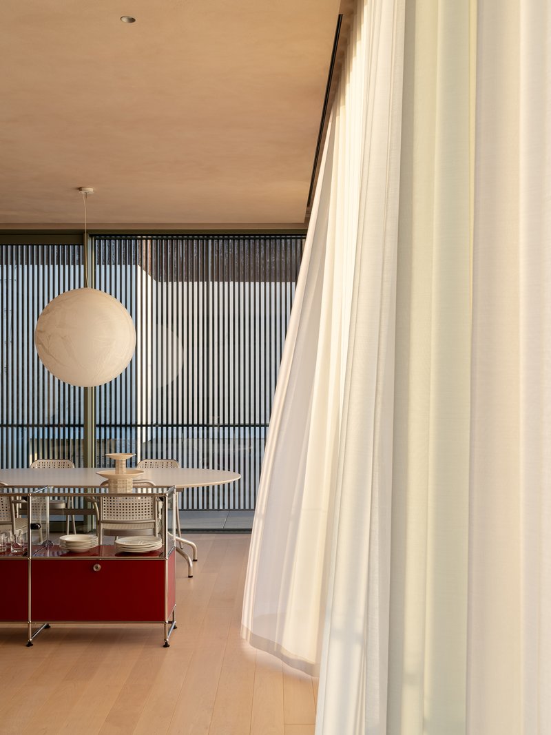 View through a translucent curtain toward vertical wood slats and a red service cart under pendant lighting