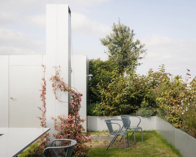 Rooftop terrace with white metal chairs and climbing vines along white walls beneath a tall chimney
