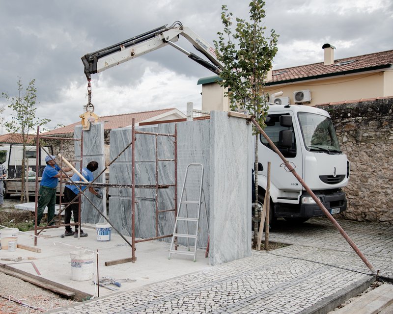 Construction workers installing prefabricated marble-clad panels with a crane-mounted truck beside young trees
