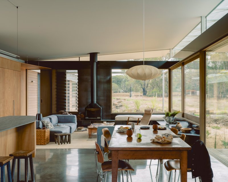 Living and dining space with freestanding wood stove and full-height glazing overlooking native landscape