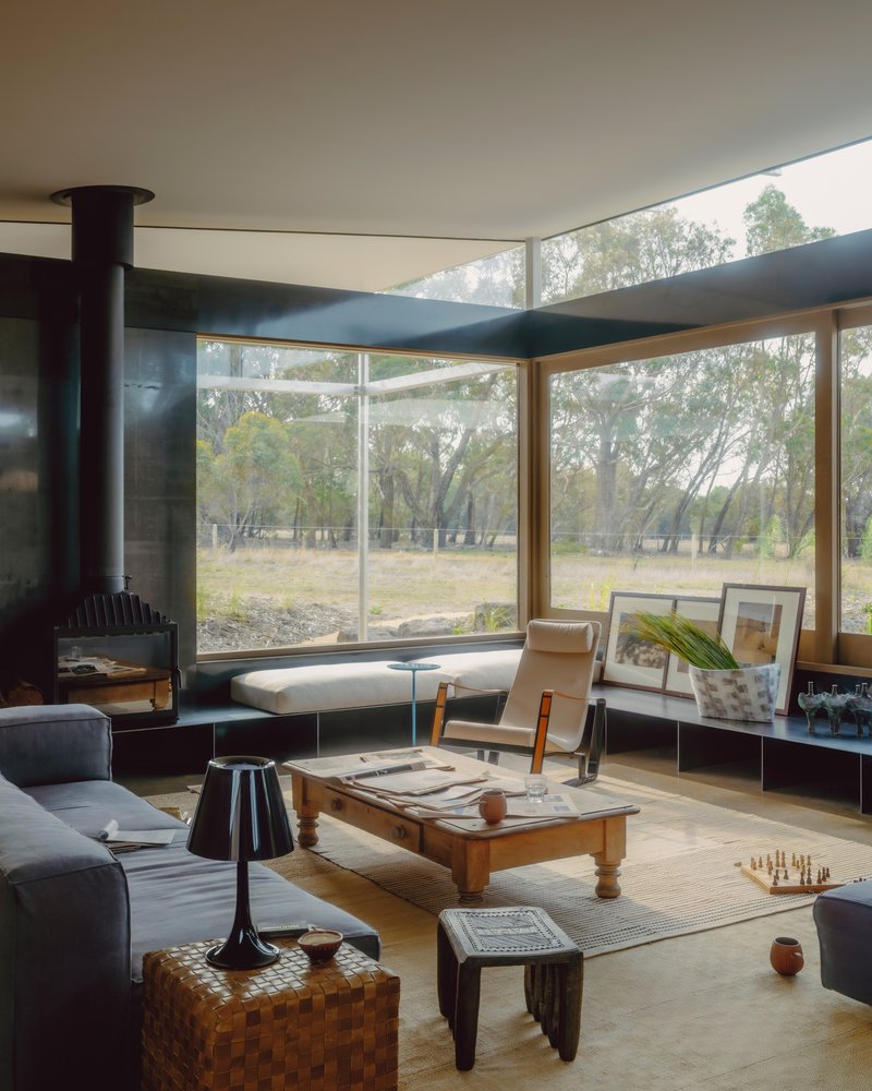 Corner seating nook with cylindrical metal fireplace beside floor-to-ceiling windows framing eucalyptus trees