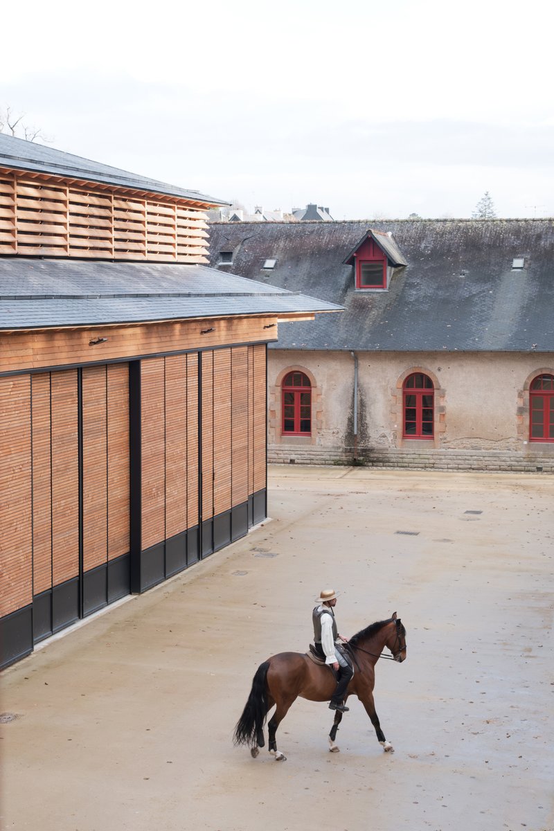 Courtyard view of the horizontal slatted timber facade beside a weathered stone stable with a rider on horseback