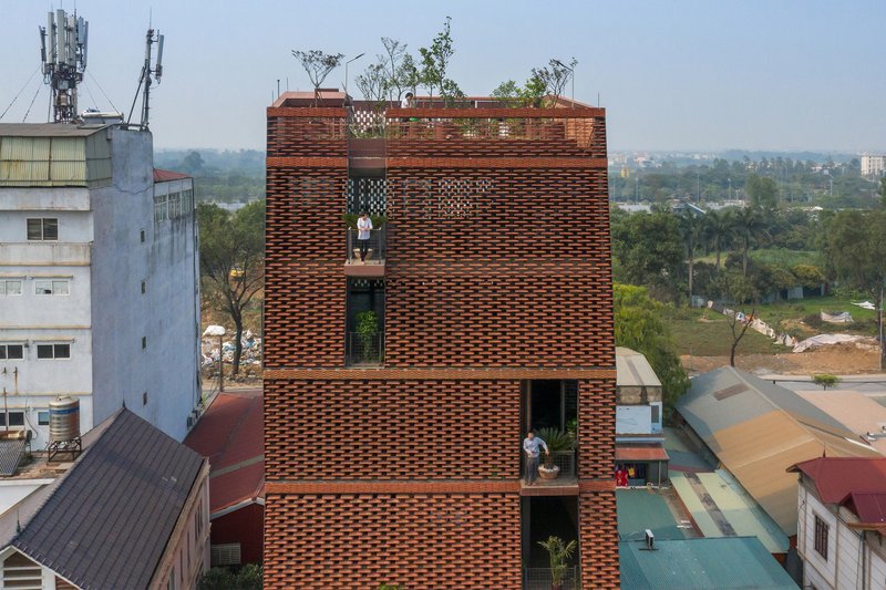 Perforated brick tower rising above neighboring rooftops with planted terraces and people visible in openings