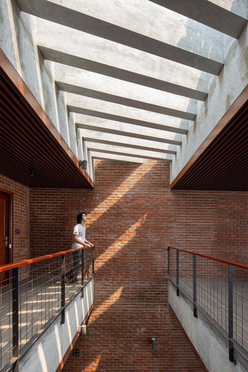 Skylit courtyard with red brick walls and concrete beams creating striped shadows on a figure at the railing