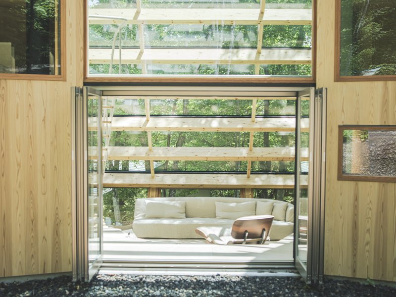 Large pivoting glass door opening from plywood-lined interior to timber-framed porch with white sofa in dappled forest light