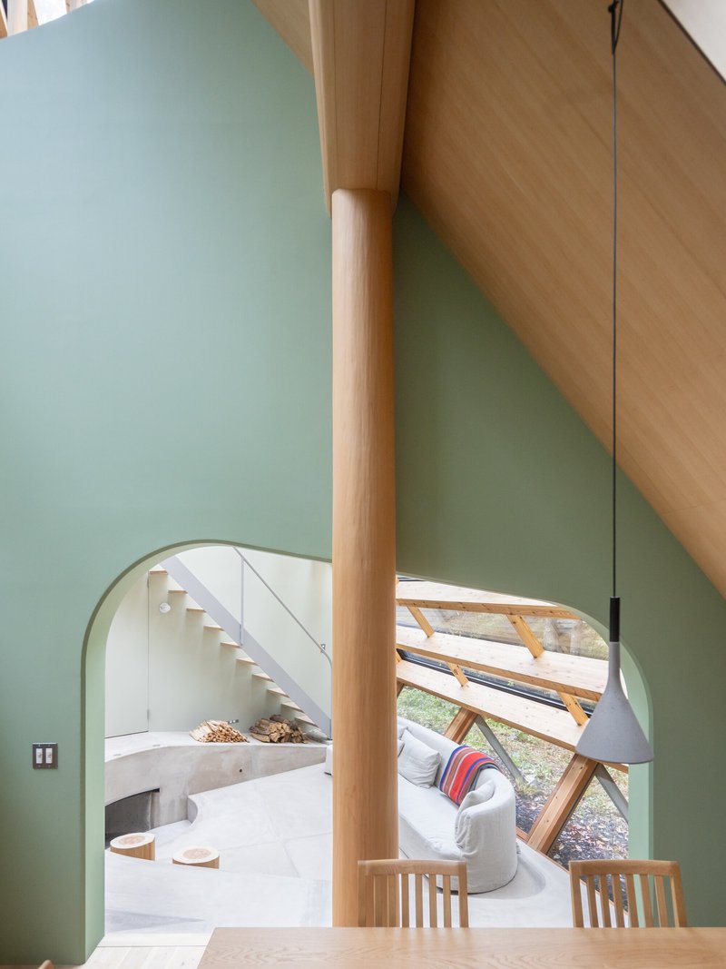 Round timber column between pale green walls with arched opening revealing white stairs and glass-walled living area beyond