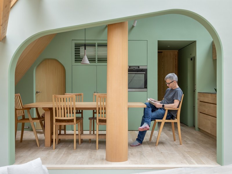 Dining area framed by pale green arched opening with resident reading at timber table beside kitchen cabinets