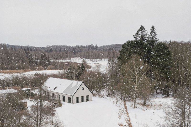 Aerial view of white gabled house nestled among bare trees in a snowy landscape during snowfall