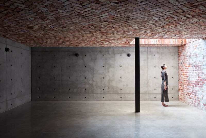 Interior space with board-formed concrete walls beneath a herringbone brick vault ceiling and a single figure