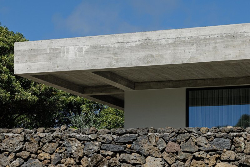 Cantilevered concrete beam with board-formed soffit above a volcanic stone retaining wall under clear sky