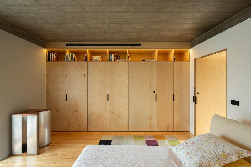 Bedroom with full-height plywood storage wall and backlit upper shelving beneath a timber ceiling