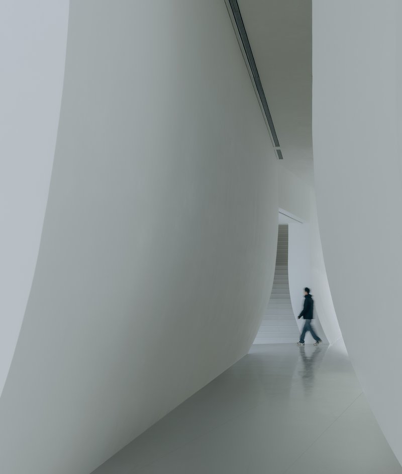 White interior corridor with a person walking beneath curving walls and a linear ceiling slot