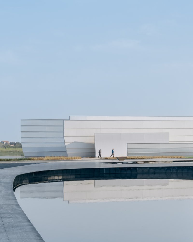 Curved water channel beside the white layered facade with two figures walking on the plaza
