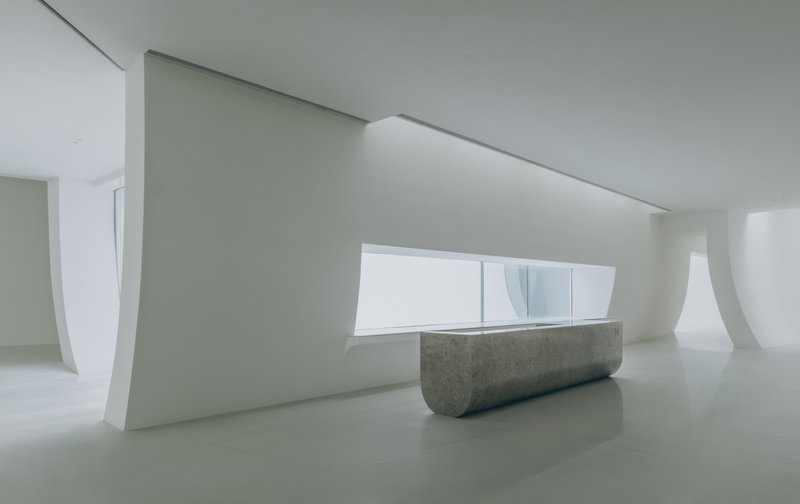Reception desk in grey concrete beneath a horizontal window framed by curved white walls