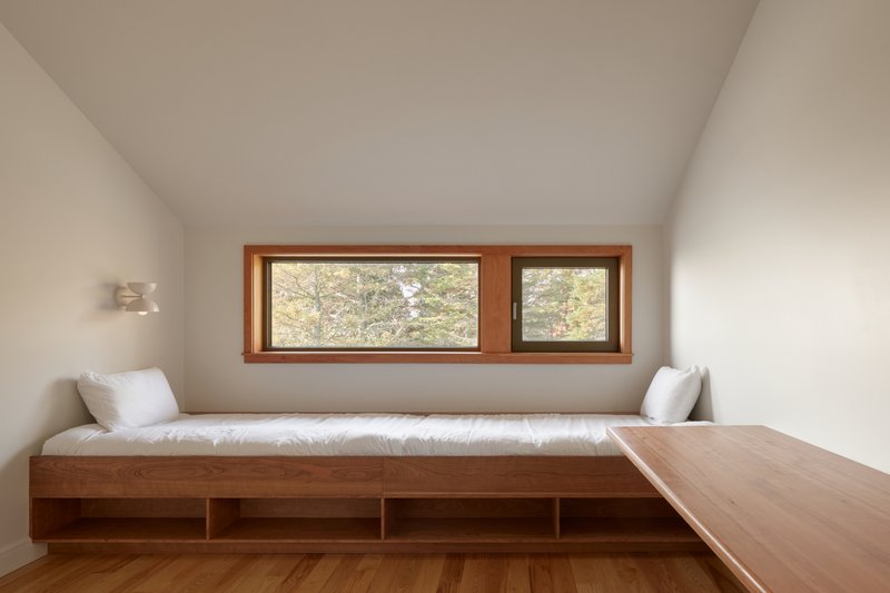 Built-in wood sleeping platform with storage cubbies beneath horizontal windows framing pine branches