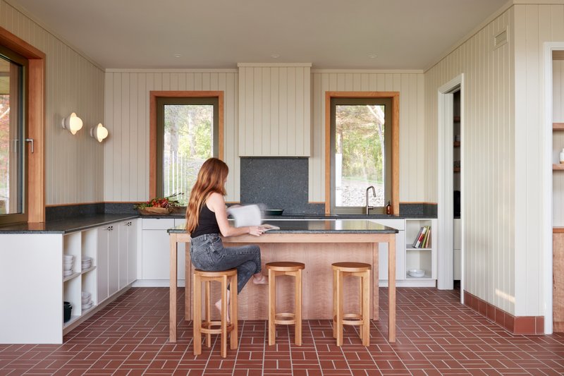 Kitchen with timber island, three stools, and a person seated reading at the counter near two windows