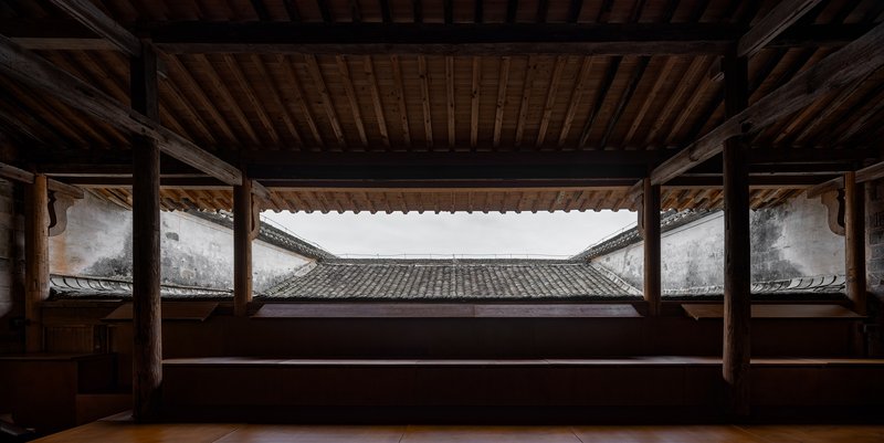 Framed view through timber structure to courtyard with tiled roof and white walls under overcast sky