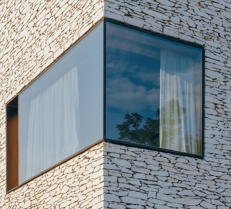 Corner window detail set into rough-textured stone cladding with sky reflection in glass
