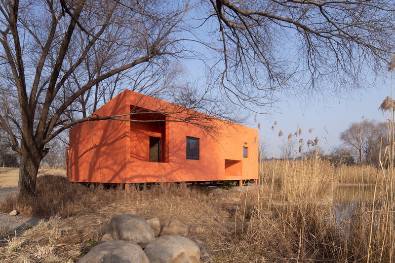 Red-orange facade with recessed openings on a raised platform among bare trees and dry vegetation
