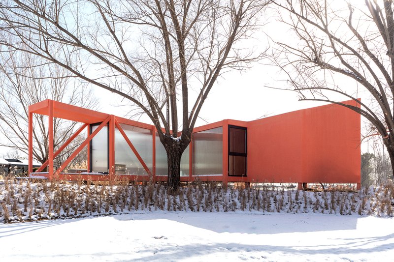 Coral-colored volume with steel-framed glazed enclosure and solid wall emerging from snow-covered ground