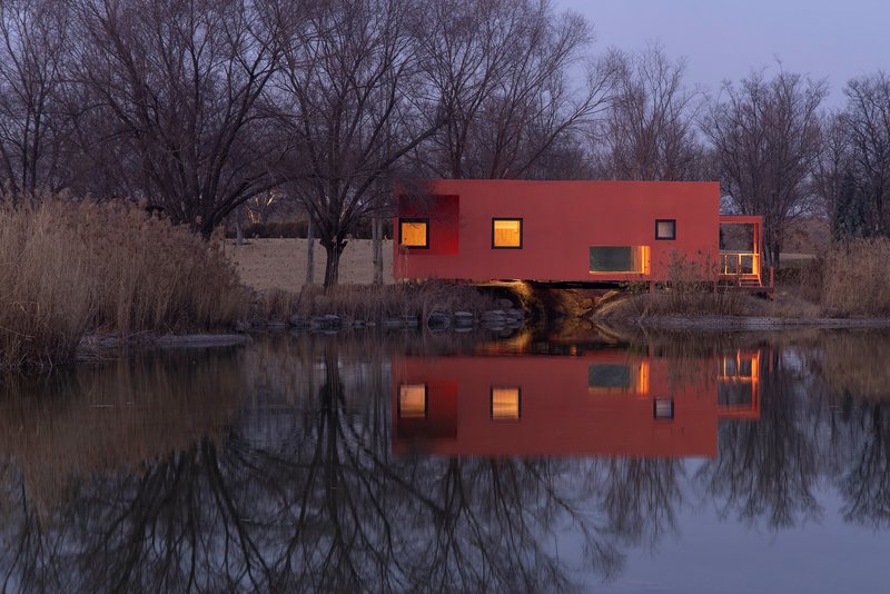 Red metal facade with illuminated square windows reflecting in pond water at dusk among bare trees