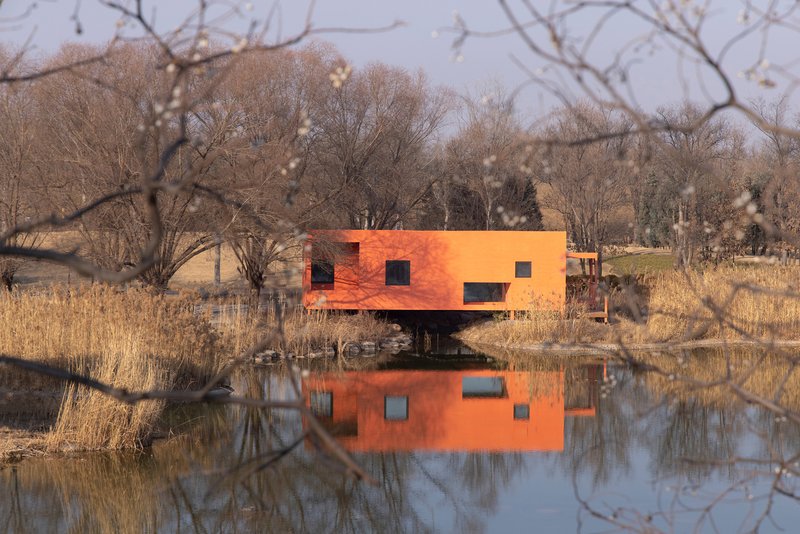 Orange metal-clad volume elevated on stilts reflected in a pond surrounded by dormant winter grasses