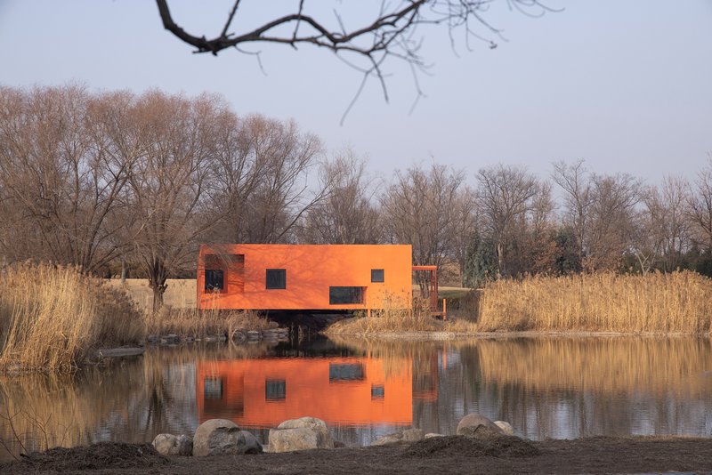 Orange facade with punched windows raised above pond on single column with reeds and rock shoreline