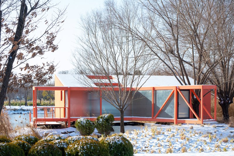 Red structural frame with glazed panels and white roof on snow-covered ground beneath bare trees