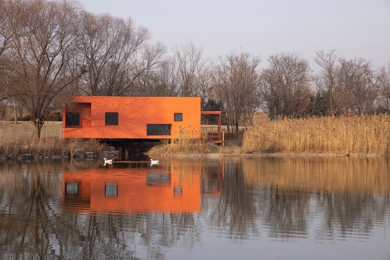 Orange metal-clad volume on stilts reflected in the pond with geese and dormant reeds