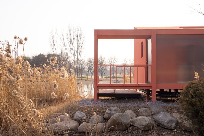 Elevated terrace with red steel frame and glass railings overlooking a pond and dormant reeds