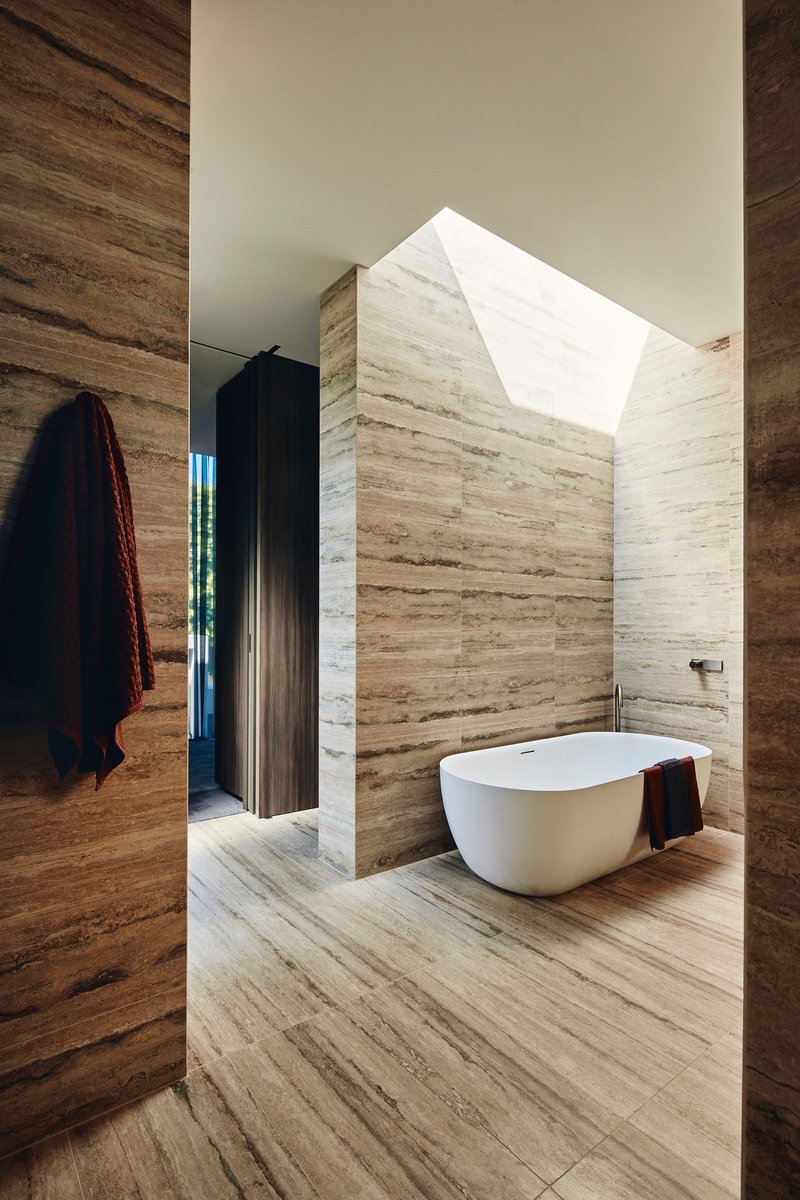 Freestanding white bathtub in travertine-clad bathroom with angled skylight overhead