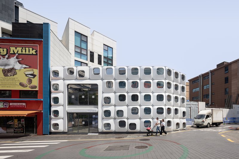 Streetscape showing the curved white facade with circular windows among neighboring buildings and passing vehicles