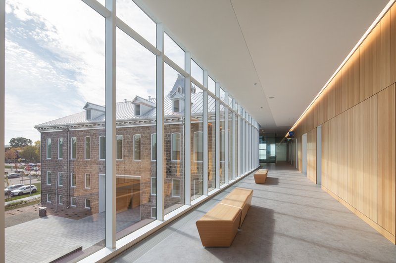 Glazed hallway with vertical white mullions framing views of the adjacent masonry building and parking area