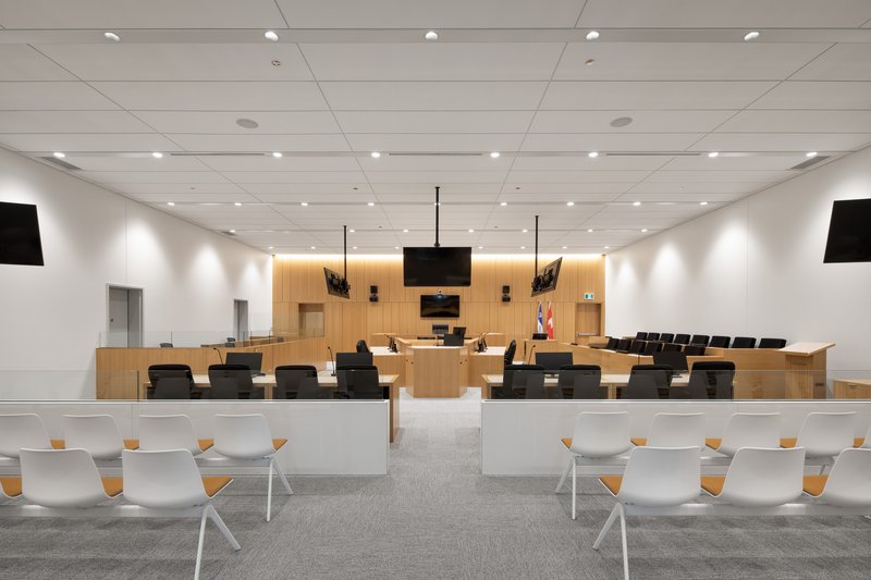 Courtroom interior with timber judicial bench, black monitors, and rows of gallery seating under recessed lighting