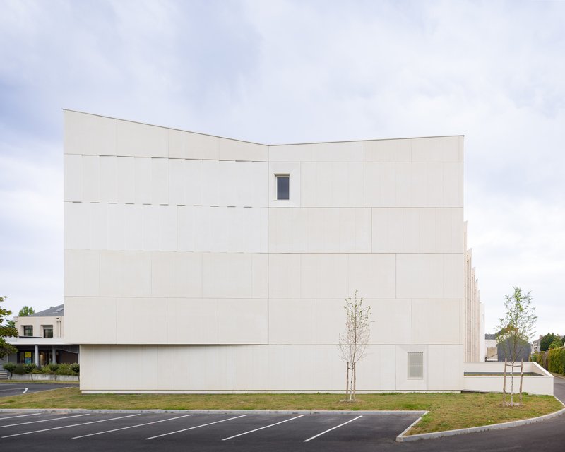 Cream-coloured precast concrete panel facade with minimal window openings flanked by young saplings and parking lot