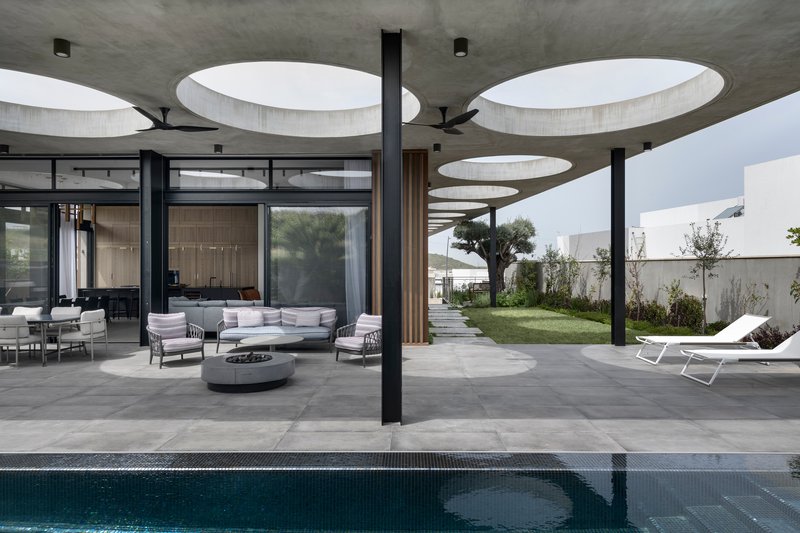Covered outdoor terrace with circular ceiling voids and black columns overlooking a pool and garden