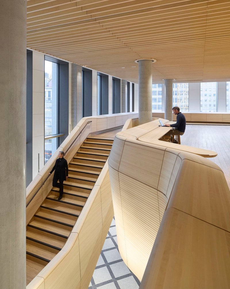 Curved bleacher seating with wood slat cladding beneath a linear wood batten ceiling as person works