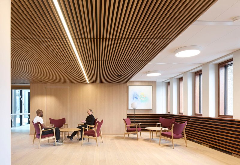 Meeting lounge with burgundy chairs beneath wood batten ceiling and circular skylights by windowed wall