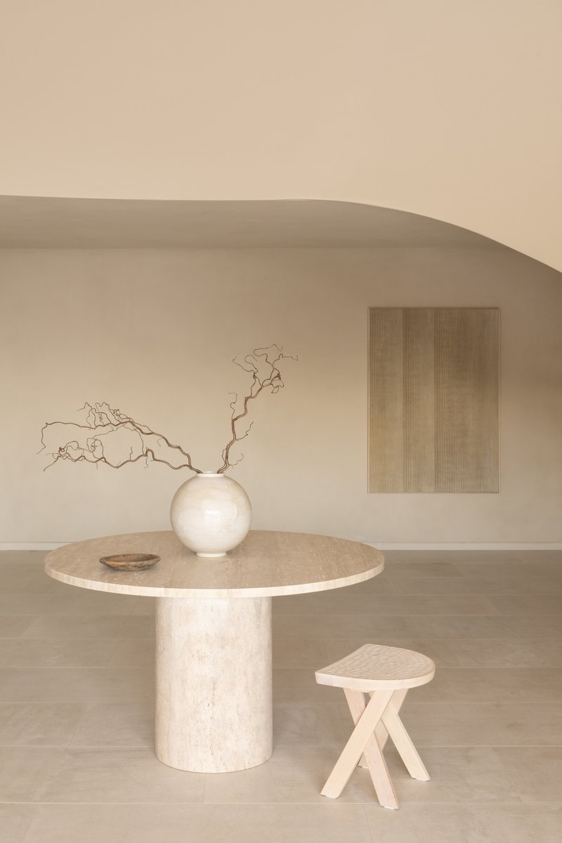 Travertine dining table with spherical vase and dried branches beneath a curved plaster ceiling