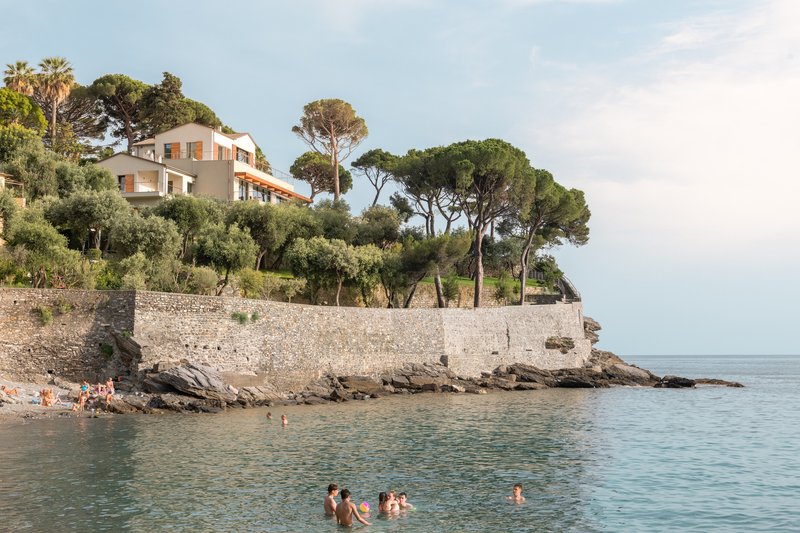 Coastal elevation set into a rocky hillside with umbrella pines and swimmers in the cove below