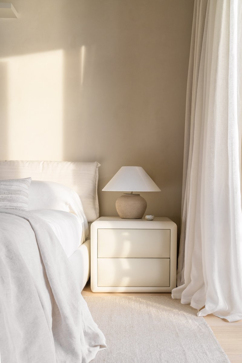 Bedside nightstand with table lamp beside white linen and floor-length curtains in soft morning light