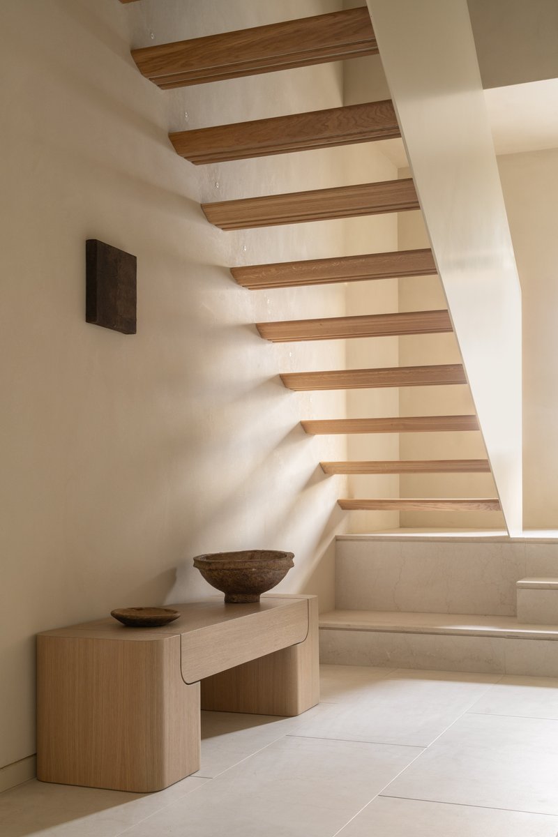 Floating timber staircase with open risers rising along a white plaster wall in warm daylight
